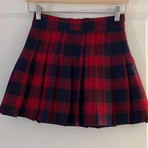 Rutig röd och marinblå plisserad kjol - Superfin kort plisserad kjol i rött och marinblått rutigt mönster från Fashion. Kjolen har bred linning och är helfodrad med svart innerfoder. Perfekt för en cool och trendig look.