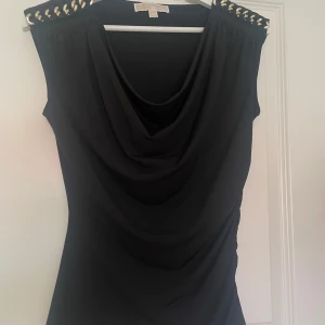 Svart draperad topp Michael Kors - Svart ärmlös topp från Michael Kors med snygg drapering framtill och guldfärgade kedjedetaljer på axlarna. Toppen har en elegant, lös passform och är tillverkad i ett mjukt, stretchigt material som känns skönt mot huden.
