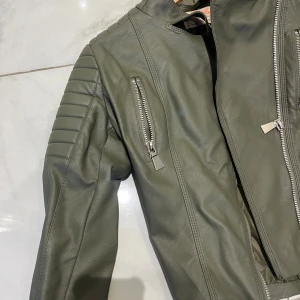Grön skinnjacka med dragkedjor - Snygg grön skinnjacka med coola detaljer som silvriga dragkedjor och vadderade partier på axlarna. Jackan har en modern bikerstil och hög krage. Perfekt för dig som vill sticka ut med en edgy look.