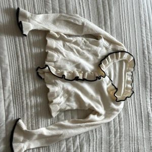 Vit kofta med volang och pärlknappar - Supersöt vit kofta med svarta kanter och volangdetaljer längs krage och ärmslut. Stängs med pärlknappar framtill. Stickad i mjukt material och har långärmad passform. Perfekt för dig som gillar en romantisk och chic stil.