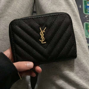 Svart quiltad plånbok med dragkedja - Säljer en svart quiltad plånbok med guldigt logotypemblem framtill. Plånboken har dragkedja runtom och flera fack på insidan för kort och sedlar. Tillverkad i strukturerat skinn med snyggt mönster och exklusiv känsla.