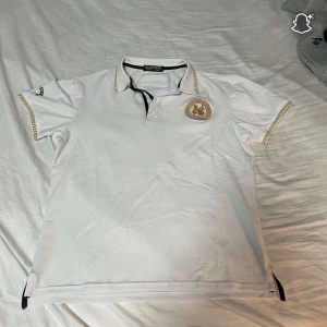 Vit polo t-shirt från Blacklisted - Vit polo t-shirt från Blacklisted med guldfärgade detaljer och mönster längs ärmarna och kragen. Har en broderad logga på bröstet och klassisk krage med knappslå. Perfekt för dig som gillar stilrena och exklusiva plagg.