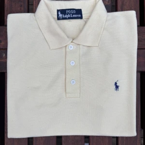 Beige Polo Ralph Lauren t-shirt - Snygg beige pikétröja från Polo Ralph Lauren med klassisk krage och tre vita knappar framtill. Broderad marinblå logga på bröstet. Tillverkad i mjuk bomull som andas. Perfekt för dig som gillar stilren och enkel design.