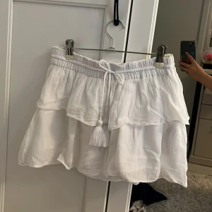 Söt vit volangkjol från Gina Tricot🫶🏻 - Superfin vit kortkjol från Gina Tricot med volanger och resår i midjan. Kjolen har knytband med pärl- och tofsdetaljer framtill och är gjord i ett lätt bomullstyg. Perfekt för sommarens alla tillfällen!