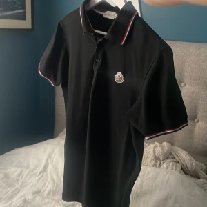 Svart Moncler pikétröja med röd/vit kant - Säljer en svart pikétröja från Moncler med korta ärmar och klassisk krage. Tröjan har röda och vita detaljer på krage och ärmslut samt Moncler-logga på bröstet. Perfekt för en clean och stilren look. Storlek L men liten i storleken så rekommenderar att man vanligtvis bär s eller m, jag är 183 och väger 82 kg så ser ni hur den sitter på mig på sista bilden.