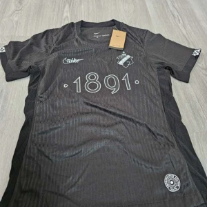 Svart AIK fotbollströja Nike 1891 - Snygg svart AIK fotbollströja från Nike med diskreta ränder, klubbmärke och årtalet 1891 på bröstet. Texten 'SMOKINGLIRARE' finns i nacken. Tröjan är kortärmad och tillverkad i ett lätt, ventilerande material som passar perfekt till match eller träning. Finns i både S och M, vid frågor är det bara att höra av sig 