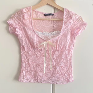 Rosa spetsblus med rosett från Dazy - Supersöt ljusrosa blus i spets från Dazy med korta ärmar, söt rosett framtill och lätt puff vid axlarna. Blusen har en romantisk vibe med transparent spets och en liten volangkant vid halsringningen. Perfekt för dig som gillar en feminin och trendig look.