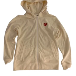 CDG Zip hoodie - Snygg beige hoodie från Comme des Garçons Play med dragkedja och huva. Tröjan har det ikoniska röda hjärtat broderat på bröstet och praktiska fickor framtill. Perfekt för en chill och stilren look. (Kan gå ner i pris vid snabb affär) 