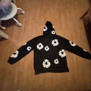 Svart hoodie med vita blommor Denim Tears - Svart hoodie från Denim Tears i storlek M, prydd med stora vita blommor över hela tröjan och huvan. Tröjan har en klassisk huva och långa ärmar, perfekt för dig som gillar streetwear med unik design. Materialet känns mjukt och bekvämt mot huden. Säljer pga att den inte var min smak. För fler frågor är det bara att skriva privat