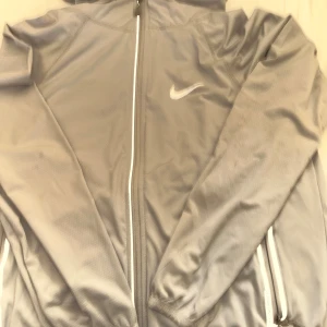Grå Nike vindjacka med huva - Perfket för nån som är över 165 till 173Snygg grå vindjacka från Nike med huva och hel dragkedja framtill. Jackan har diskret struktur i tyget, två fickor med dragkedja och klassisk Nike-logga på bröstet. Perfekt för dig som gillar sportig och clean stil. 