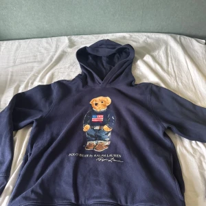 Mörkblå hoodie Polo Bear Ralph Lauren - Säljer en mörkblå hoodie från Polo Ralph Lauren med ikonisk Polo Bear-tryck på bröstet. Tröjan har huva och långa ärmar, tillverkad i mjuk bomull. Perfekt för dig som gillar klassisk streetstyle och vill sticka ut med en cool björn på framsidan.