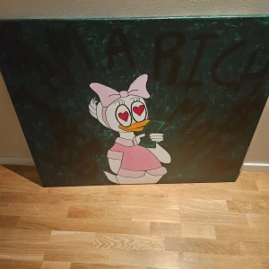 Tavla  - Det här är en färgstark tavla med motiv av Kajsa Anka, klädd i rosa och med hjärtformade ögon, som håller i pengar. Bakgrunden har texten 'I AM RICH' i stora bokstäver. Perfekt för dig som gillar popkultur och vill ha något unikt på väggen.
