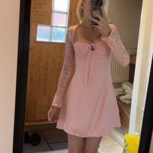 Rosa spetsklänning med snörning - Supersöt ljusrosa kortklänning med långa spetsärmar och snörning både fram och bak. Klänningen har en feminin v-ringning och romantiska detaljer som ger en riktigt gullig vibe. Perfekt för dig som gillar en söt och trendig look. Snörning som en korsett i ryggen, snörning i nacken och snörning vid bysten, klänningen har även en dragkedja i sidan. 