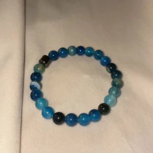 Blått pärlarmband med stenar - Snyggt armband med runda pärlor i olika blå nyanser och en unik svart kubformad pärla. Elastiskt band gör det enkelt att ta på och av. Perfekt accessoar för att ge din outfit lite extra färg och stil.