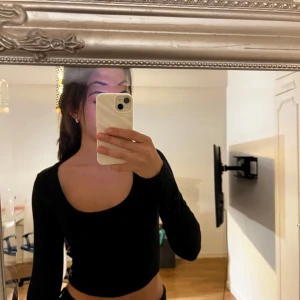 Svart croppad långärmad topp - Säljer en svart croppad topp med lång ärm och rundad, djup urringning. Materialet är ribbat och stretchigt, vilket gör den både bekväm och snygg. Perfekt att styla med höga jeans eller kjol för en trendig look.