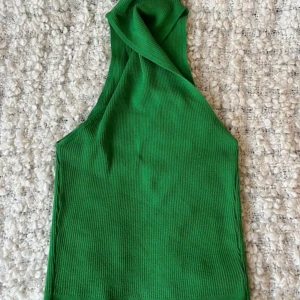 Grön ribbad halternecktopp - Säljer en snygg grön ribbad topp med halternack-design. Toppen är ärmlös och har en cool asymmetrisk halsringning. Kan användas både bak & fram så man antingen har twist-designen eller den platta delen fram.