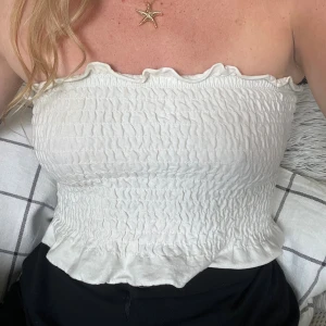 Vit bandeau tubtopp med volangkant - Supersöt vit bandeau tubtopp med smockad struktur och volangkant upptill och nedtill. Toppen är ärmlös och har en stretchig passform som sitter snyggt på kroppen. Perfekt för varma dagar och enkel att styla med jeans eller kjol. Vet tyvärr varken storlek eller varumärke då jag klippte bort lapparna av bekvämlighetsskäl, men gissar stl S men den är väldigt stretchig så passar nog XS-M.