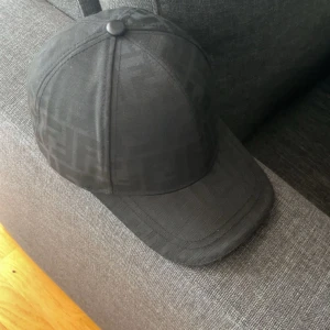 Svart Fendi keps med mönster - Snygg svart keps från Fendi med diskret FF-mönster över hela kepsen. Justerbar rem bak i svart skinn med Fendi-logga. Klassisk böjd skärm och stilren look som lyfter din outfit. Perfekt för dig som gillar designer och streetstyle.