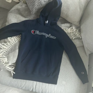 Mörkblå Champion hoodie med logga - Säljer en mörkblå hoodie från Champion med stor broderad logga över bröstet. Klassisk känguruficka framtill, huva och långa ärmar. Perfekt för chill dagar och enkel att matcha med jeans eller joggers.