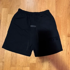 Svarta Essentials Fear of God shorts - Svarta shorts från Essentials Fear of God med stilren logga på benet och snörning i midjan. Modellen är loose fit och har elastisk midja för skön passform. Materialet är mjuk bomull, perfekt för chill dagar och streetwear vibes.