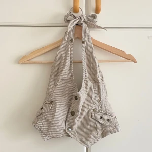 Randig halterneck korsett-topp - Säljer en beige och vit randig halterneck korsett-topp med knyt i nacken och snygga knappar framtill. Toppen har två fickdetaljer med knappar och en croppad passform. Perfekt för dig som vill ha en unik och trendig look i sommar. Toppen är intagen midjan från en storlek m till s