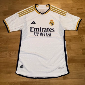 Real Madrid player version 23/24 hemmatröja L - Säljer en officiell Real Madrid hemmatröja. Den är äkta och har alla originaldetaljer, inklusive klubbmärke, Adidas-logga och sponsortryck på framsidan. Materialet är Adidas HEAT.RDY, vilket gör den extra lätt, luftig och bekväm. Det är perfekt både till träning, match eller bara som en snygg vardagströja. Storleken är L men modellen är slim fit, så den kan även passa dig som brukar ha M och vill ha den lite mer normal i passform. Skicket är mycket bra då den är knappt använd. 