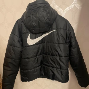 Svart pufferjacka från Nike Therma-Fit - Säljer en svart pufferjacka från Nike Therma-Fit med stor vit logga på ryggen och mindre logga på bröstet. Jackan har huva, dragkedja framtill och två fickor. Perfekt för kyliga dagar och riktigt snygg streetstil. Nypris 700kr