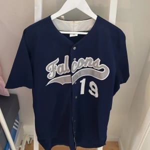 Blå Falcons Baseballtröja Wilson - Snygg blå baseballtröja från Wilson med Falcons-tryck och siffran 19 i vitt och silver på både fram- och baksida. Klassisk basebollinspirerad design med knappar framtill och korta ärmar. Perfekt för dig som gillar sportig stil.