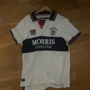 Vit Morris Cricket Club t-shirt - Vit t-shirt från Morris Cricket Club med mörkblå bred rand över bröstet och tryck. Klassisk krage med röd, vit och blå kant, samt korta ärmar med matchande detaljer. Snygg broderad logga och patch på bröstet. Perfekt för dig som gillar sportig stil. Sitter mer som en m.