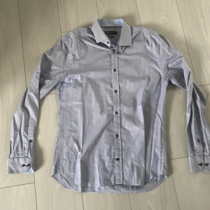 Slim fit rutig skjorta från Riley - Snygg slim fit skjorta från Riley i blåvitt smått rutigt mönster. Skjortan har långa ärmar, klassisk krage och bruna knappar. Perfekt för dig som gillar stilrena och fräscha plagg. Tillverkad i bomull för en skön känsla hela dagen. Prkekt skick 