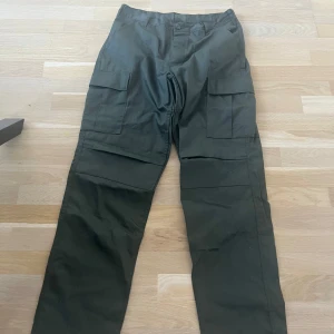 Mörkgröna cargopants med fickor - Säljer ett par mörkgröna cargopants med flera stora fickor på sidorna och bak. Byxorna har rak passform och är tillverkade i slitstarkt bomullstyg. Midjan är justerbar mellan 31 och 35 tum och innerbenslängden är 29,5 till 32,5 tum.
