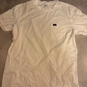Vit t-shirt från Lee med ficka - Vit t-shirt från Lee i mjuk bomull, klassisk rund hals och korta ärmar. På bröstet finns en praktisk ficka med en liten svart Lee-logga. Enkel och clean design som funkar till jeans eller shorts. Perfekt för dig som gillar stilrena plagg.