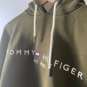 Olivgrön hoodie från Tommy Hilfiger - Säljer en olivgrön hoodie från Tommy Hilfiger med broderad logga och text på bröstet. Tröjan har huva med vita snören och en stor magficka. Perfekt för dig som gillar streetwear och vill ha något bekvämt och snyggt magficka är pyttelite avslitet