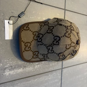 keps med GG-mönster och läderdetalj - Snygg keps i beige och brunt med det klassiska GG-monogrammet över hela kepsen. Justerbar rem baktill i brunt skinn och detaljer i samma färg. Kepsen är tillverkad i bomull och polyester, perfekt för dig som vill sticka ut med en lyxig accessoar. Köpt av en kompis och har inte eller något bevis därför pris