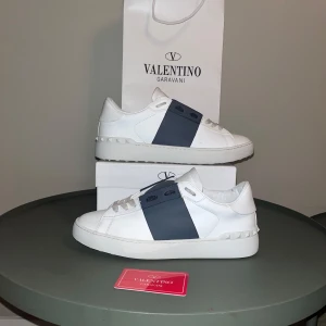 Valentino Garavani Open sneakers vit/blå - Snygga Valentino Garavani Open sneakers i vitt skinn med bred blå rand över mitten. Klassisk låg modell med rund tå, vita snören och diskreta nitar på hälen. Perfekta för dig som gillar stilrena och exklusiva sneakers med en twist.