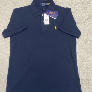 Marinblå Polo Ralph Lauren t-shirt - Snygg marinblå t-shirt från Polo Ralph Lauren i slim fit-modell. Klassisk piké med krage, två knappar och den ikoniska gula polospelaren broderad på bröstet. Tillverkad i mjuk bomull som känns skön mot huden. Perfekt för dig som gillar stilren och sportig look.