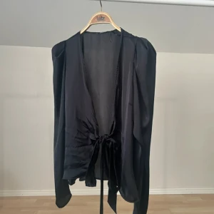 Svart omlottblus med knyt - Säljer en svart omlottblus i glansigt, tunt material med långa ärmar och v-ringning. Blusen har en snygg knytning framtill som ger en fin look. Perfekt att styla med jeans eller kjol. 