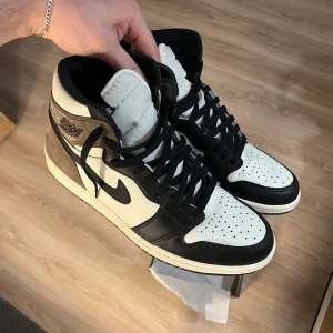 Nike Air Jordan 1 High Mocha - Nike Air Jordan 1 High Mocha med svart och vit läder, bruna mockadetaljer och klassisk svart swoosh. Storlek 47. Höga sneakers med snörning, perforerad tå och ikonisk Jordan-logga på sidan. Perfekt för dig som vill sticka ut med en clean och tidlös look.