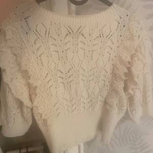 Vit stickad tröja med volangdetaljer - Supersöt vit stickad tröja med vackert hålmönster och breda volangdetaljer längs ärmarna. Tröjan har rund halsringning och långa ärmar, perfekt för dig som gillar romantisk och feminin stil.
