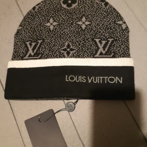 Svart och vit Louis Vuitton mössa - Snygg svart och vit mössa från Louis Vuitton med klassiskt LV-monogram och logga framtill. Mössan har en bred vit kant och är stickad i mjukt material. Perfekt accessoar för att lyfta din vinterstil.