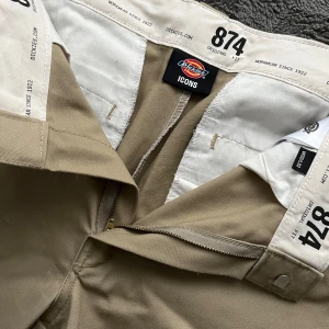 Beiga Dickies 874 byxor - Såå snygga beiga Dickies 874 byxor med straight fit och normal passform. Byxorna har hällor för bälte, dragkedja och knapp framtill samt två bakfickor. Jättefina att vika ner i midjan och ta dem från hödmidjade till lågmidjade. Har tyvärr några få hål längs ner men inget märkvärdigt. Vid frågor hör av dig!