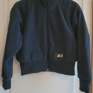 Mörkblå fleecejacka från Adidas - Säljer en mörkblå fleecejacka från Adidas med hel dragkedja framtill och ribbade muddar. Jackan har en mjuk och fluffig yta, perfekt för kyliga dagar. Adidas-logga på vänster sida. Snygg och sportig stil som funkar till många outfits.
