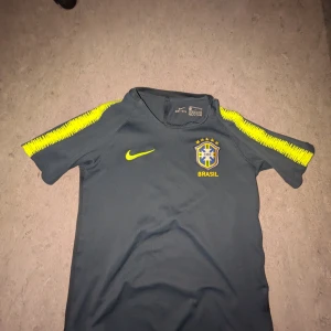 Nike Brasil fotbollströja mörkblå - Snygg mörkblå Brasilien fotbollströja från Nike med gula detaljer och Brasil-logga på bröstet. Tröjan har korta ärmar med gula mönster längs axlarna och är tillverkad i ett lätt och ventilerande material som passar perfekt till träning eller match.