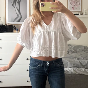 Vit blus med spets från Pigalle - Supersöt vit blus från Pigalle med v-ringning, vida trekvartsärmar och fina spetsdetaljer både framtill och på ärmarna. Blusen är croppad och har en luftig passform, perfekt till jeans eller kjol för en fräsch look.