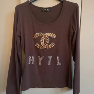 Brun långärmad tröja med pärlor - Snygg brun långärmad tröja från Y.R.U.S med rund halsringning. Framsidan har ett stort pärlbroderat motiv och texten HYTL i glittriga stenar. Perfekt för dig som gillar unika detaljer och vill sticka ut lite extra.