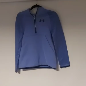 Blå hoodie från Under Armour ColdGear - Säljer en blå hoodie från Under Armour ColdGear med halv dragkedja och huva. Tröjan har en loose passform, långärmade ärmar och en stor ficka framtill. Perfekt för kyliga dagar och träning. Insidan är mjuk och fleecefodrad för extra värme.
