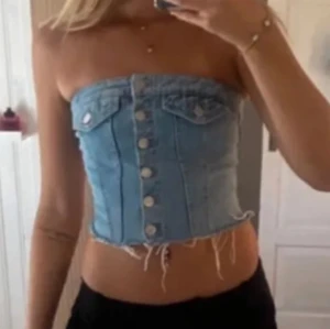 Ljusblå jeans bandeau tubtopp - Säljer en ljusblå bandeau tubtopp i jeans med knappar framtill och rå, fransig nederkant. Toppen har dekorativa fickor upptill och en tight passform som framhäver midjan. Perfekt för dig som vill sticka ut med en edgy denimlook.