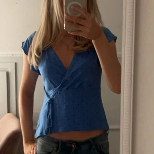 Blå omlottopp med prickar - Supersöt blå omlottopp med svarta prickar och knyt i sidan. Toppen har korta ärmar och v-ringning som ger en snygg siluett. Perfekt till jeans eller kjol för en chill och trendig look.