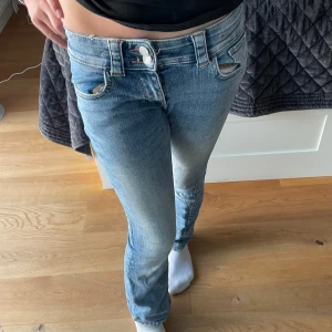 Lågmidjade bootcut jeans i ljusblå denim - Snygga lågmidjade jeans i ljusblå denim med bootcut passform. Jeansen har slitna detaljer, dubbla knappar i midjan och klassiska fickor både fram och bak med lock och knapp bak. Perfekta för en avslappnad och trendig look.