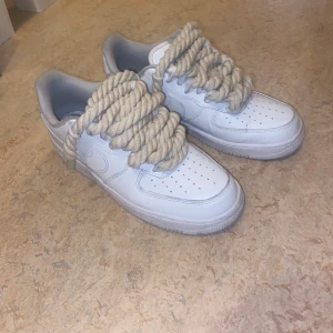 Nike Air Force 1 med rep-snören - Unika vita Nike Air Force 1 sneakers med grova, beige rep som snörning. Klassisk låg modell i skinn med perforerad tå och rund sula. Snygg och clean siluett med en twist som sticker ut i mängden.